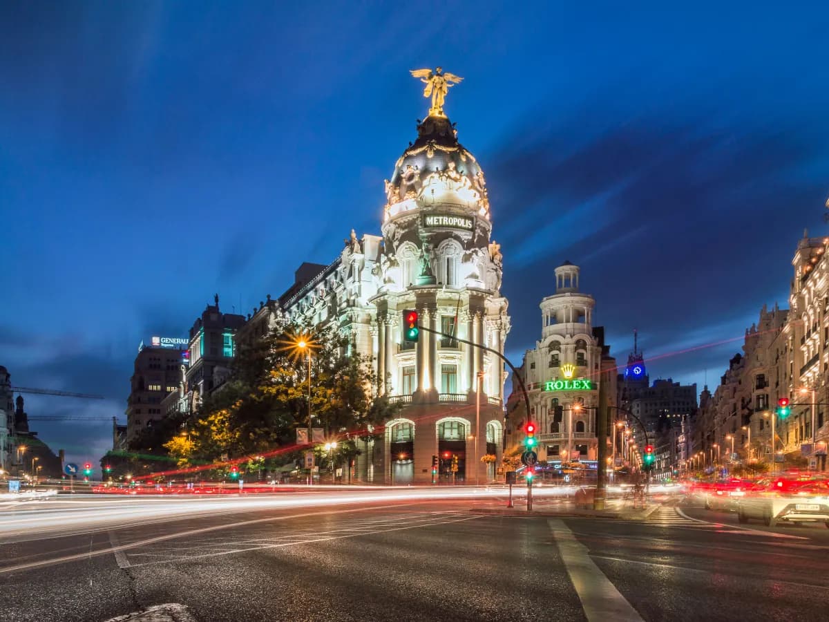 The Metropolis building lit up at dusk on Gran Via in central Madrid, with traffic light trails along the avenue.