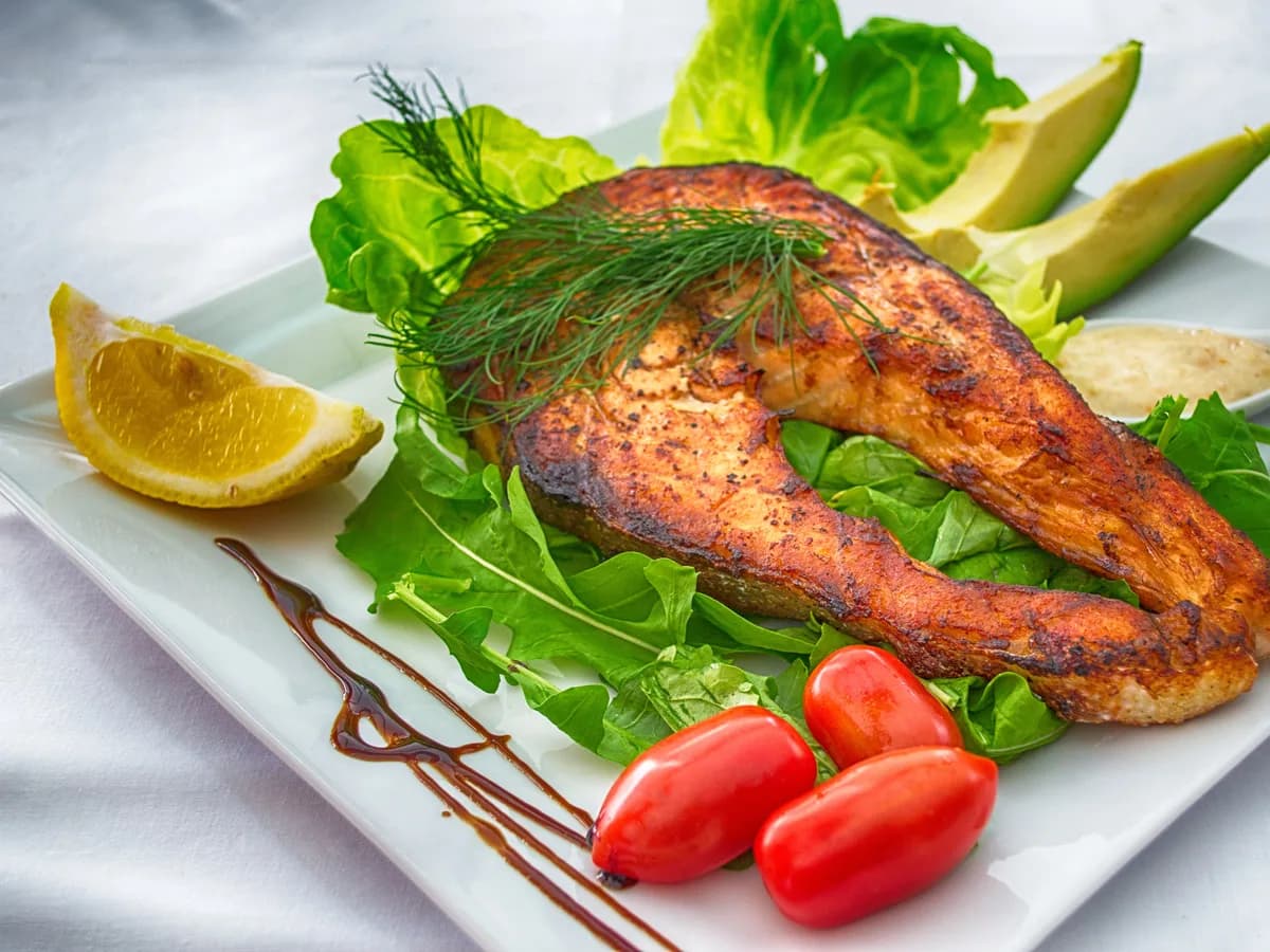 Vista cenital de un filete de salmón a la plancha con verduras frescas, aguacate, limón y tomates en un plato blanco.