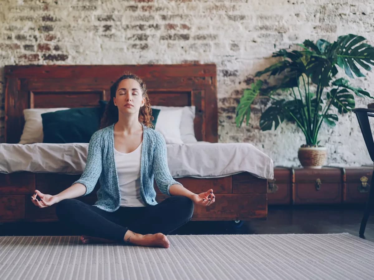 A woman sits cross-legged on the floor of a bright room, meditating with her eyes closed and hands resting on her knees.
