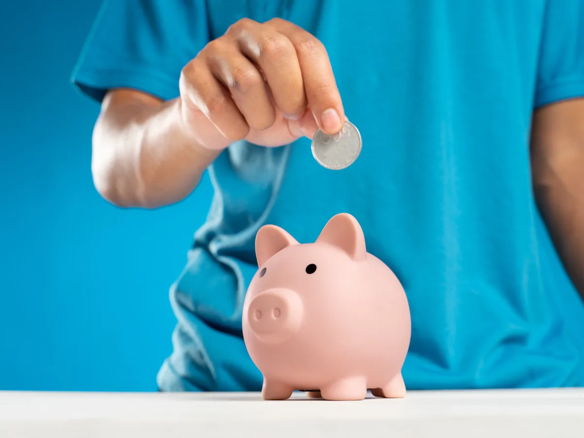 Una mano metiendo una moneda en una hucha de cerdito sobre una mesa.
