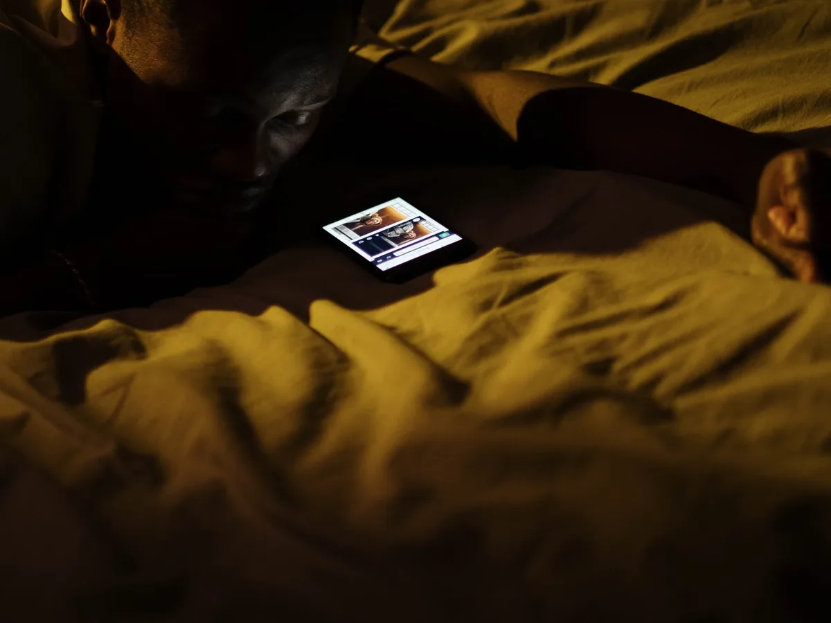 A man lying in a dark bedroom at night, his face lit by the screen of the phone in front of him.